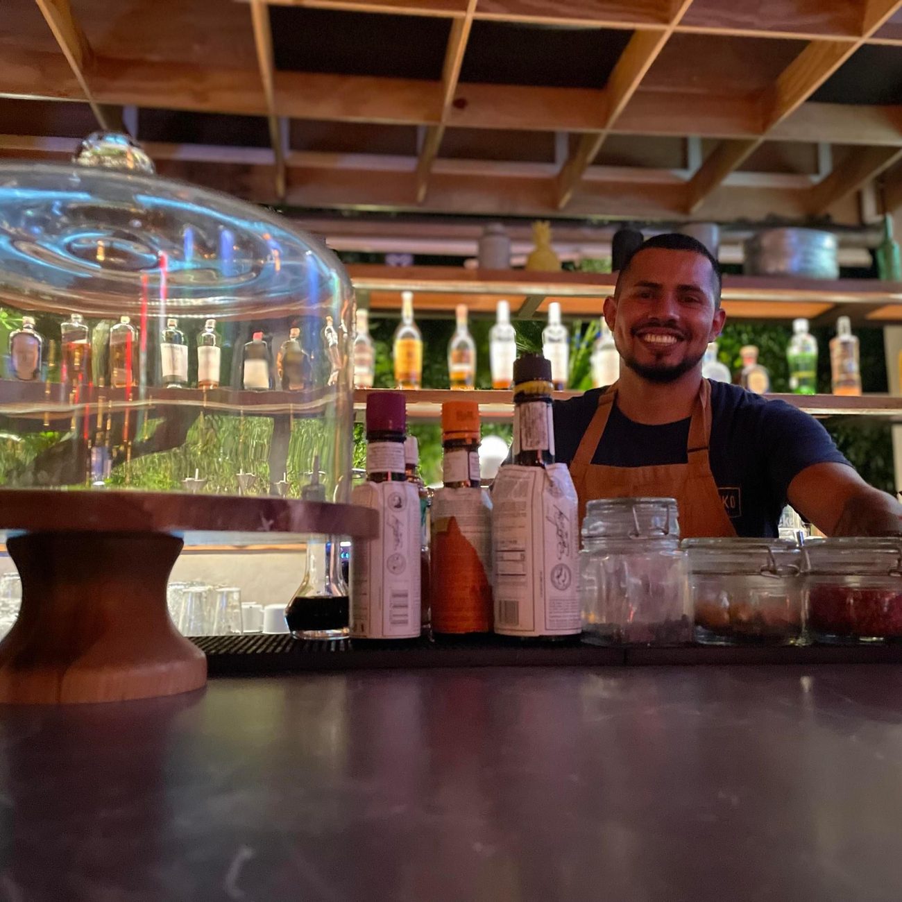 Isma smiling behind a Caribbean-style bar under a glass cloche