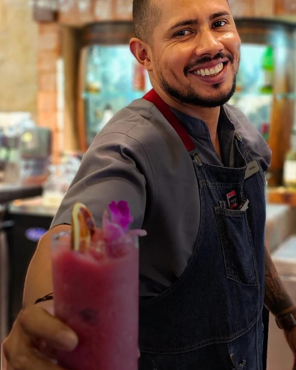 Isma smiling behind the bar, holding one of his signature flower cocktails